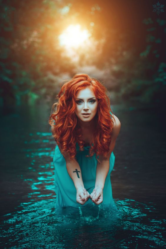 redhead beautiful enchantress in woods with corss on right arm, coming out of pond with sun setting behind her.